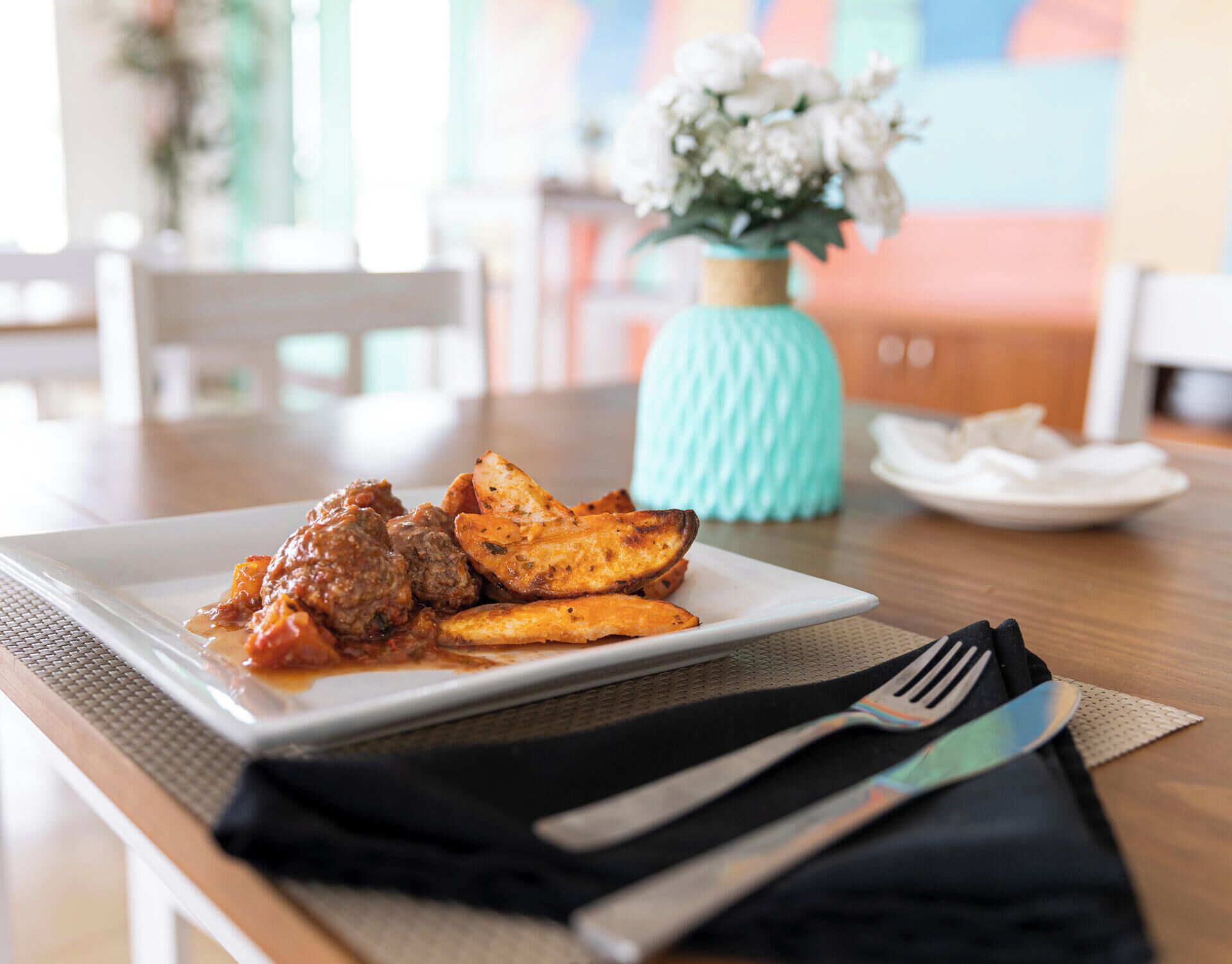 A plate of food with meatballs and roasted potatoes on a white plate, set on a wooden table with a napkin and cutlery.