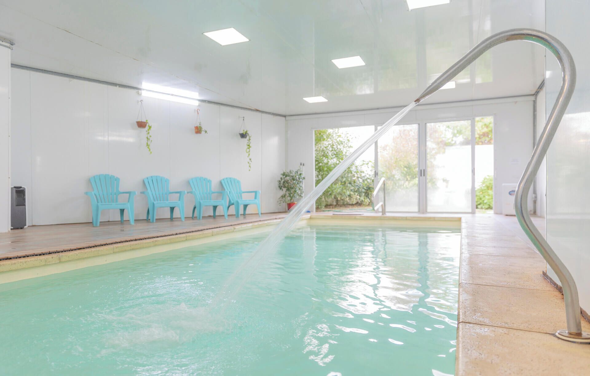Una piscina interior con vista al agua cristalina, una escalera de metal y varias sillas azules a lo largo de la pared.