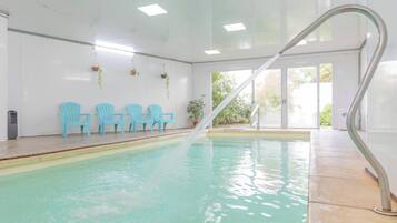Una piscina interior con vista al agua cristalina, una escalera de metal y varias sillas azules a lo largo de la pared.