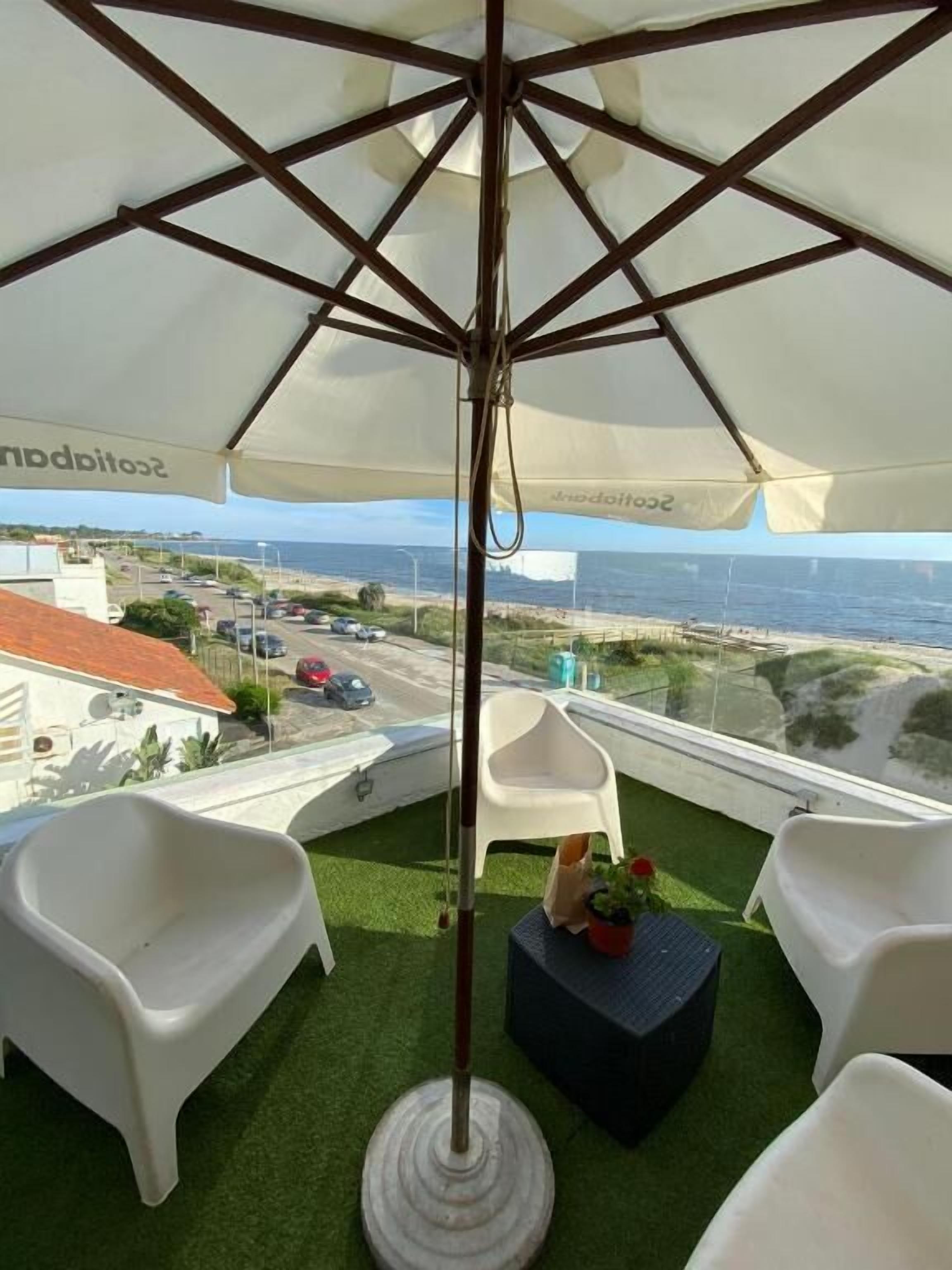 A balcony with a white umbrella, white plastic chairs, and a small table with a potted plant.