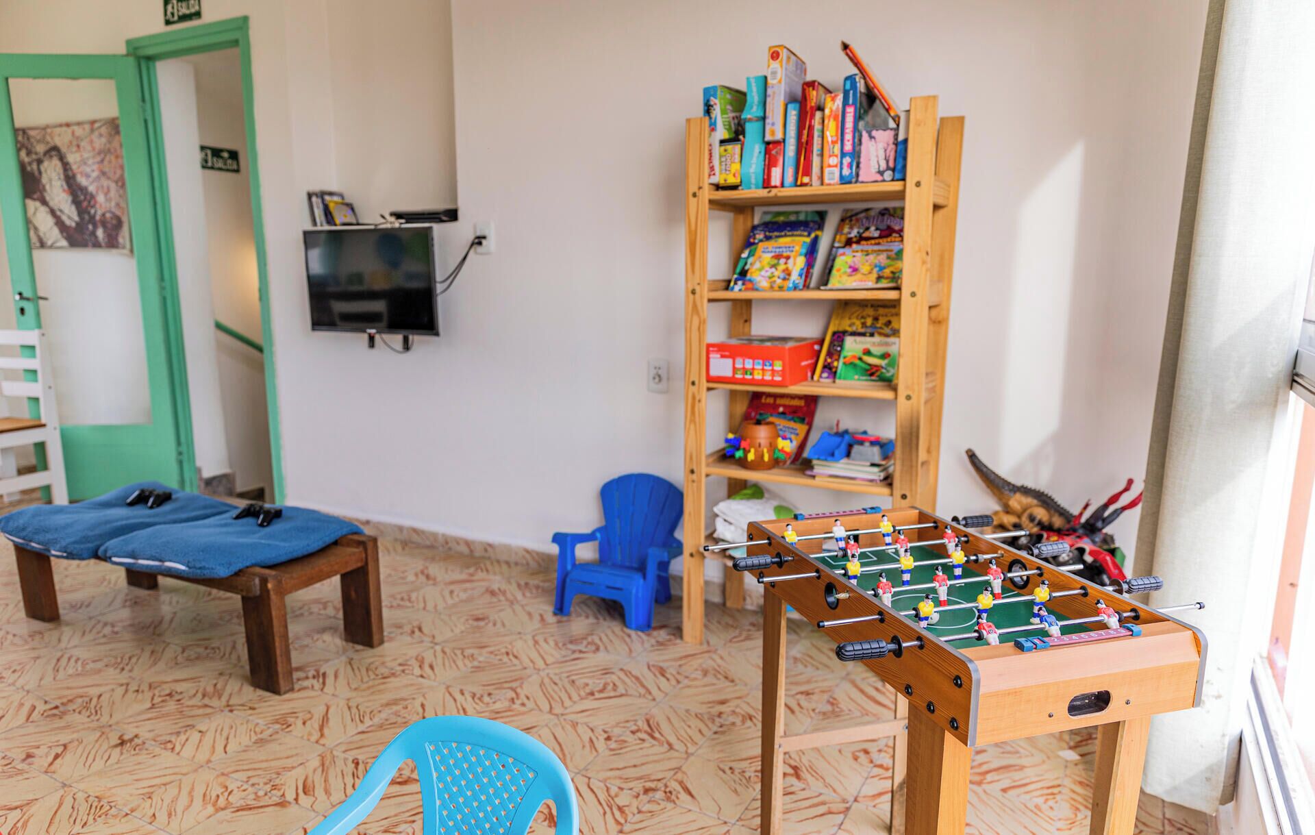 A room with a foosball table, a blue chair, a bookshelf, and a television mounted on the wall.