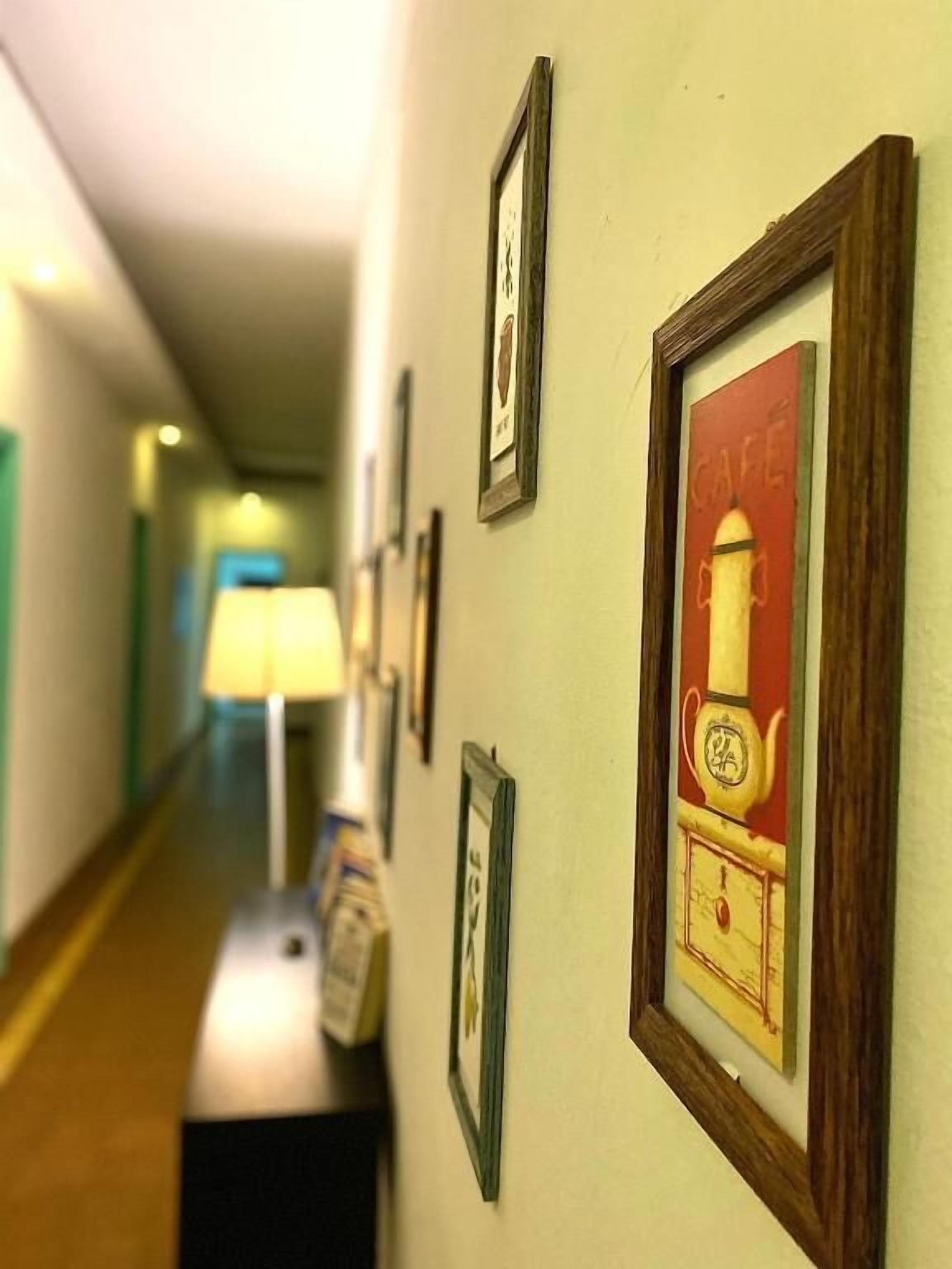 A hallway with framed pictures on the wall, a lamp, and a trash bin.