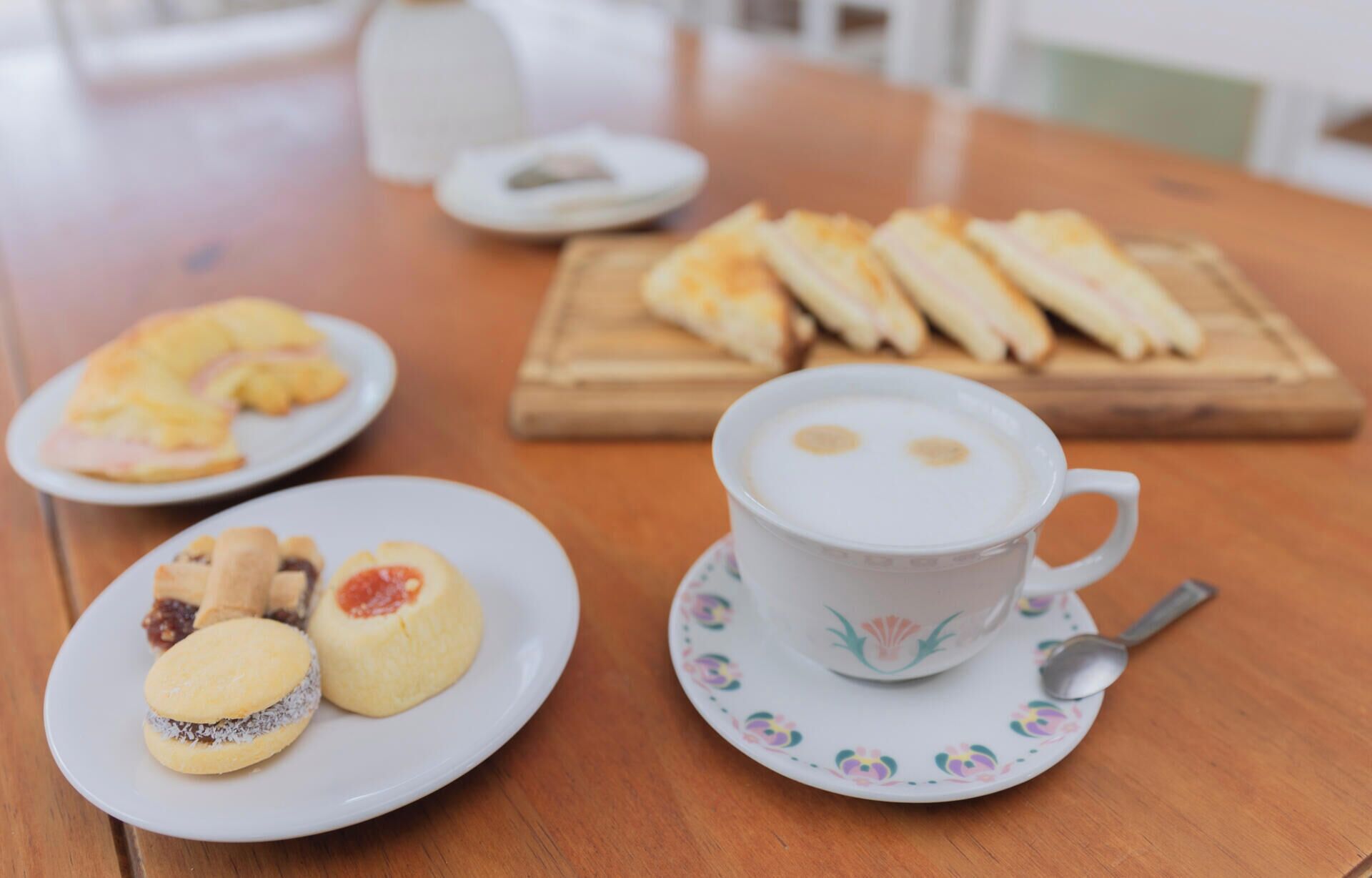 Uma mesa com uma xícara de café, um prato com biscoitos e uma tábua de madeira com doces.