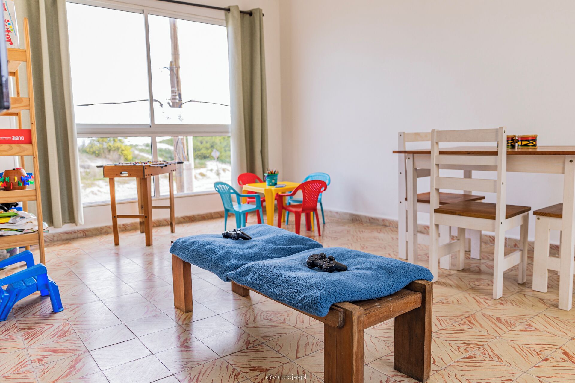 Une pièce avec un banc en bois, une petite table, des chaises colorées et une fenêtre avec des rideaux.
