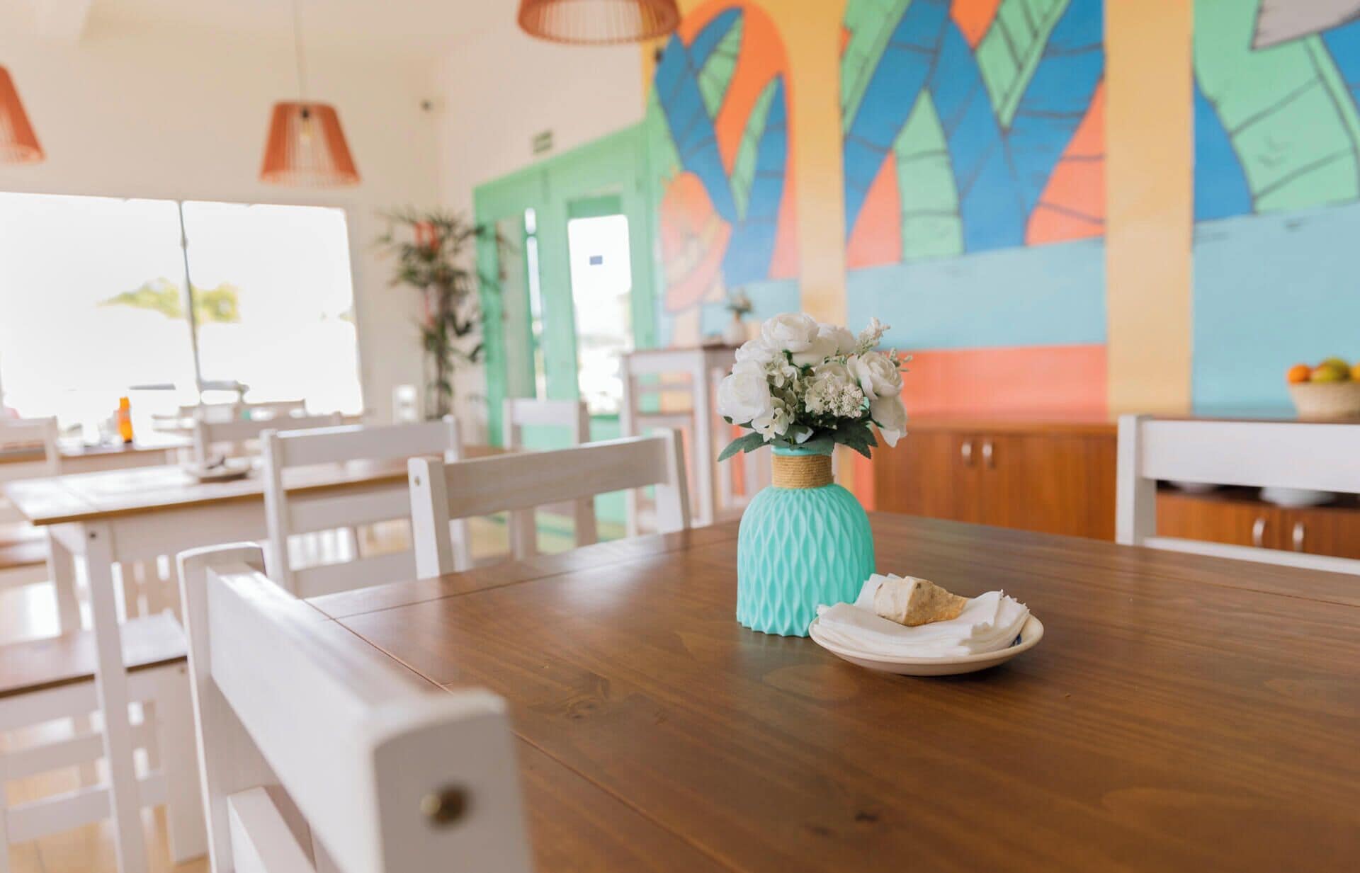 A dining table with a vase of flowers and a plate of food, set in a room with a colorful abstract wall mural.