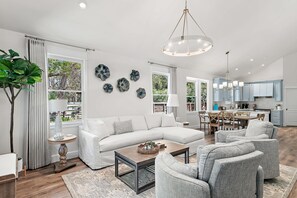 A modern living room with a white sofa, grey armchairs, and a coffee table. There is a dining area with a wooden table and chairs, and a kitchen with blue cabinets. - Seagrove Gardens - Community Pool & 4 Bikes! (Santa Rosa Beach)