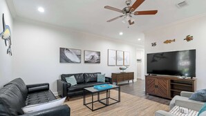 A modern living room with a black leather sofa, a wooden coffee table, a grey armchair, a TV, and a ceiling fan. - Pet-Friendly 30A Cottage w/ Fenced Yard! (Santa Rosa Beach)