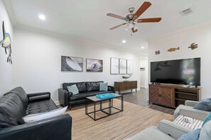 Ein modernes Wohnzimmer mit einem schwarzen Ledersofa, einem Holztisch, einem grauen Sessel, einem Fernseher und einem Deckenventilator.