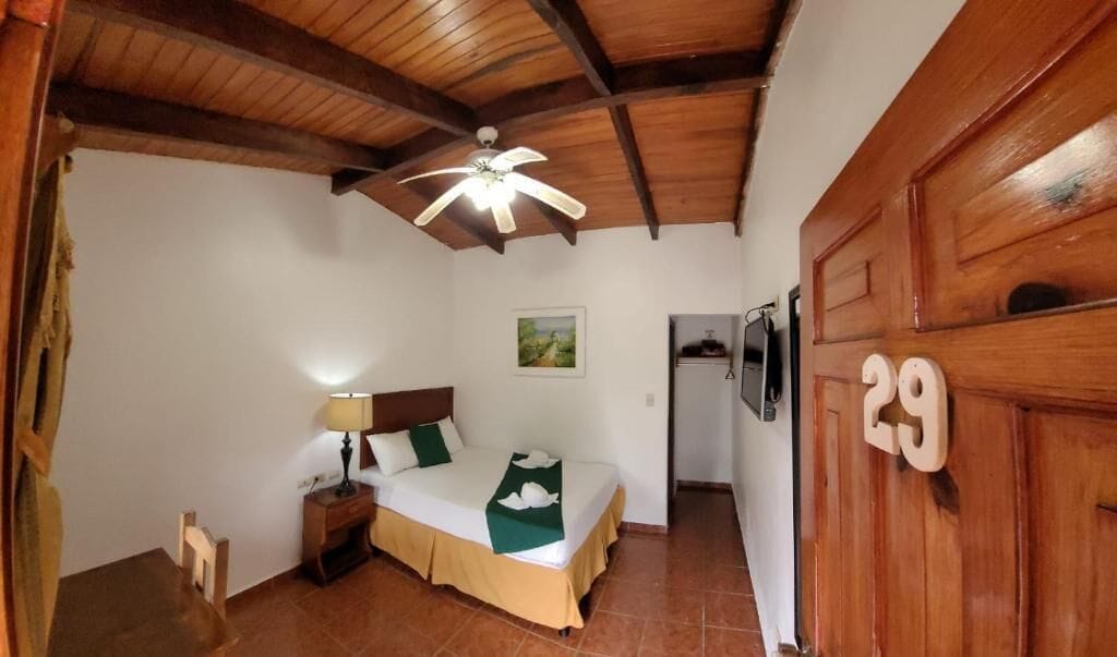 Een kamer met een bed, een nachtkastje, een lamp, een plafondventilator en een houten deur met het nummer 29.