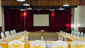 Un salón de conferencias con mesas dispuestas para una reunión, sillas con cintas amarillas, una pantalla de proyector y un telón rojo de fondo.