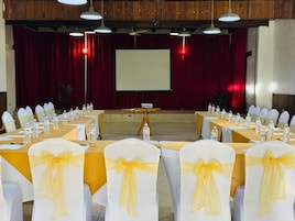 A conference room with tables set for a meeting, chairs with yellow sashes, a projector screen, and a red curtain backdrop.