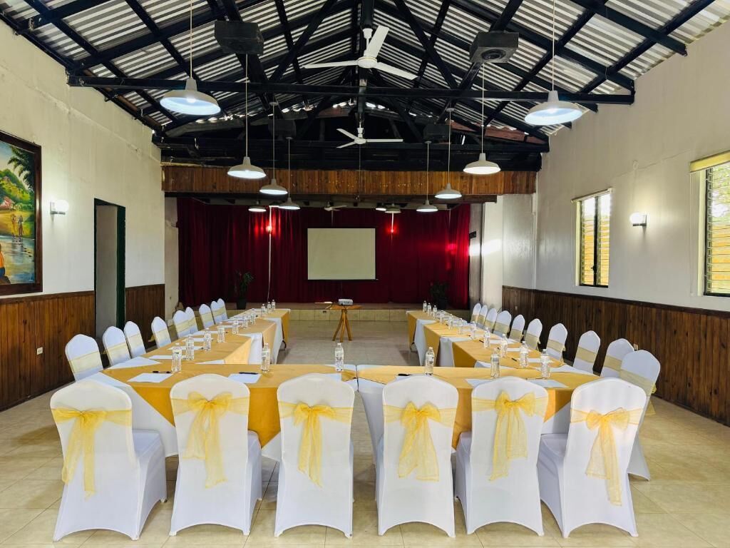Un salón de conferencias con mesas largas, sillas blancas con cintas amarillas, un escenario con cortina roja y una pantalla de proyector.