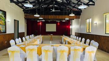 Un salón de conferencias con mesas largas, sillas blancas con cintas amarillas, un escenario con cortina roja y una pantalla de proyector.