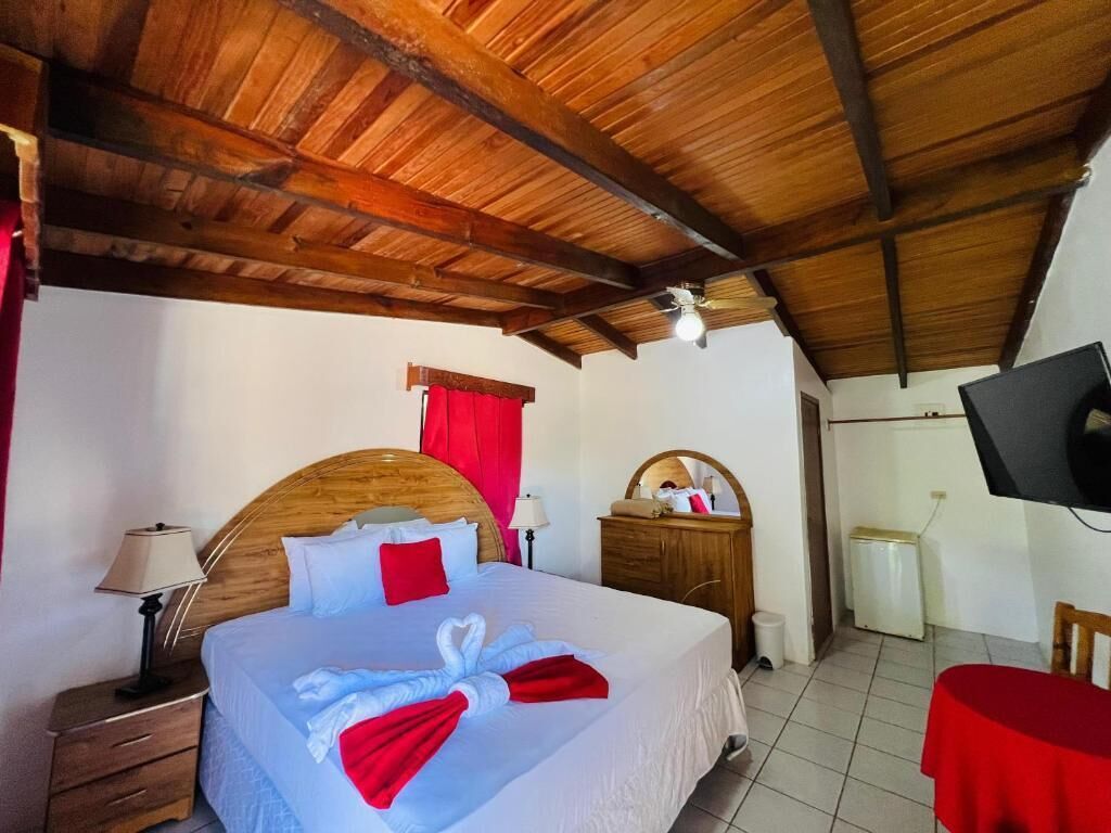 Habitación de hotel con techo de madera, cama con ropa de cama blanca y roja, mesita de noche con lámpara, cómoda de madera, televisor en la pared y una mesa roja.