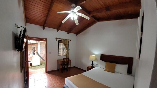 Un dormitorio con cama, escritorio, silla, ventilador de techo y ventana con cortinas.
