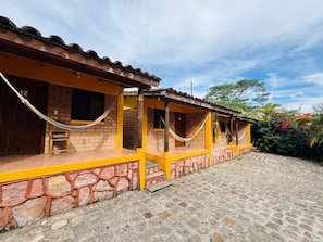 Un edificio con base de piedra y paredes pintadas de amarillo, que cuenta con un porche y una hamaca.