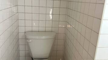 A small, white-tiled bathroom with a toilet, a trash bin, and a toilet paper holder.