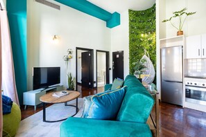 A modern living room with a teal sofa, a flat-screen TV, and a kitchen area with a refrigerator and stove. - Boutique Hotel-Style Apartment in Cape Town City (Cape Town)