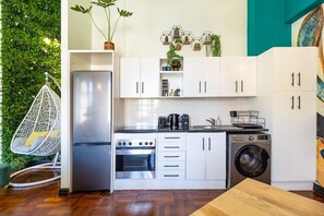Uma cozinha compacta com armários brancos, geladeira de aço inoxidável, uma cadeira suspensa e uma máquina de lavar.