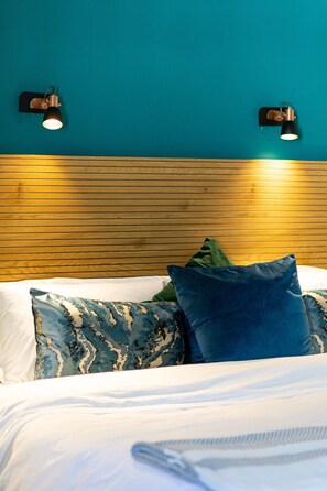 Cama com lençóis brancos e duas almofadas, uma estampada em azul e outra lisa na cor azul, em frente a uma parede azul-turquesa com duas luminárias de parede.