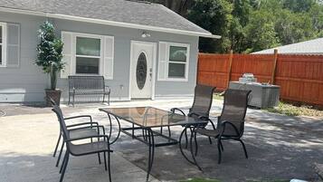 A patio with a glass-top table and four chairs, a small potted plant, and a wooden fence in the background.
