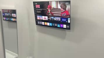 A bedroom with a bed, a TV mounted on the wall displaying a streaming service interface, and a mirror reflecting part of the room.