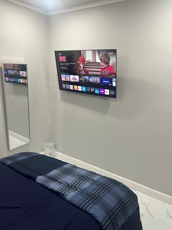 Una camera da letto con un letto, una TV a muro che mostra l'interfaccia di un servizio di streaming e uno specchio che riflette una parte della stanza.