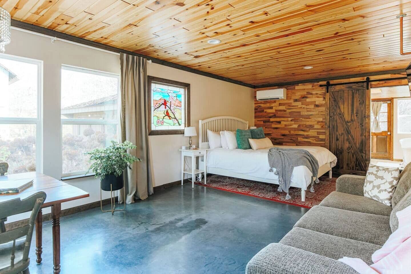 Ein Zimmer mit einem Bett, einem Schreibtisch, einem Sessel, einer Pflanze und einem Fenster mit Vorhängen.