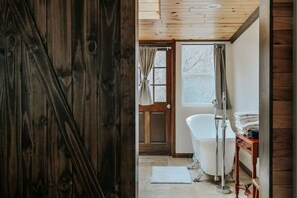 A bathroom with a white bathtub, a wooden door, and a window with curtains.