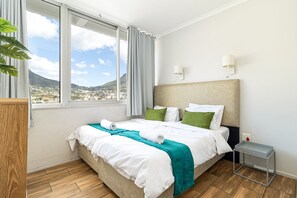 Ett hotellrum med en säng, ett nattduksbord, en lampa och utsikt över en stadssiluett genom stora fönster.