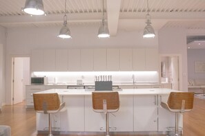 A modern kitchen with white cabinets, a central island with bar stools, pendant lighting, and a visible microwave. - Cape Colosseum Luxury Stay (Cape Town)