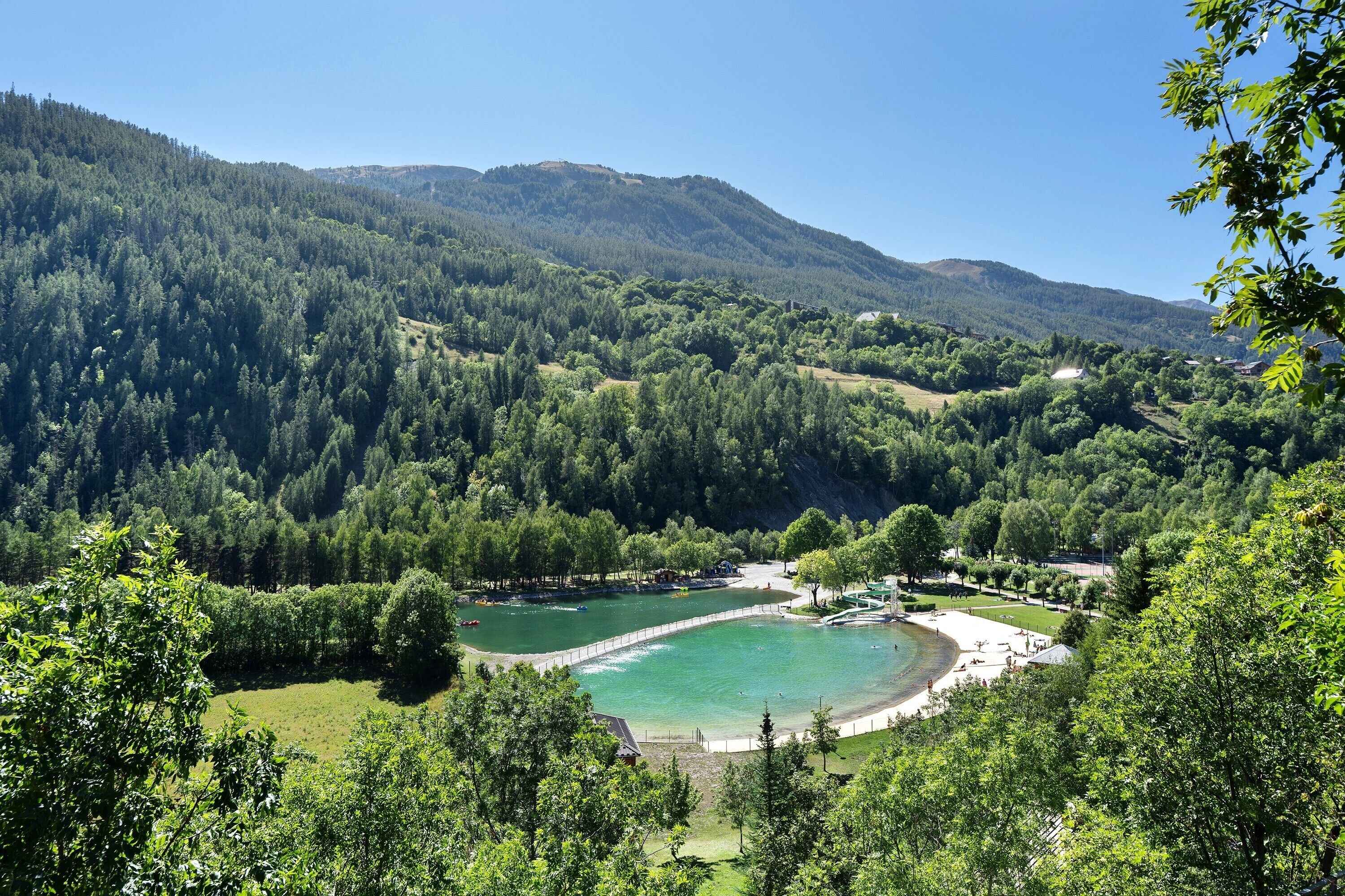 A mountainous landscape with a clear lake, surrounded by dense forests and a few buildings.