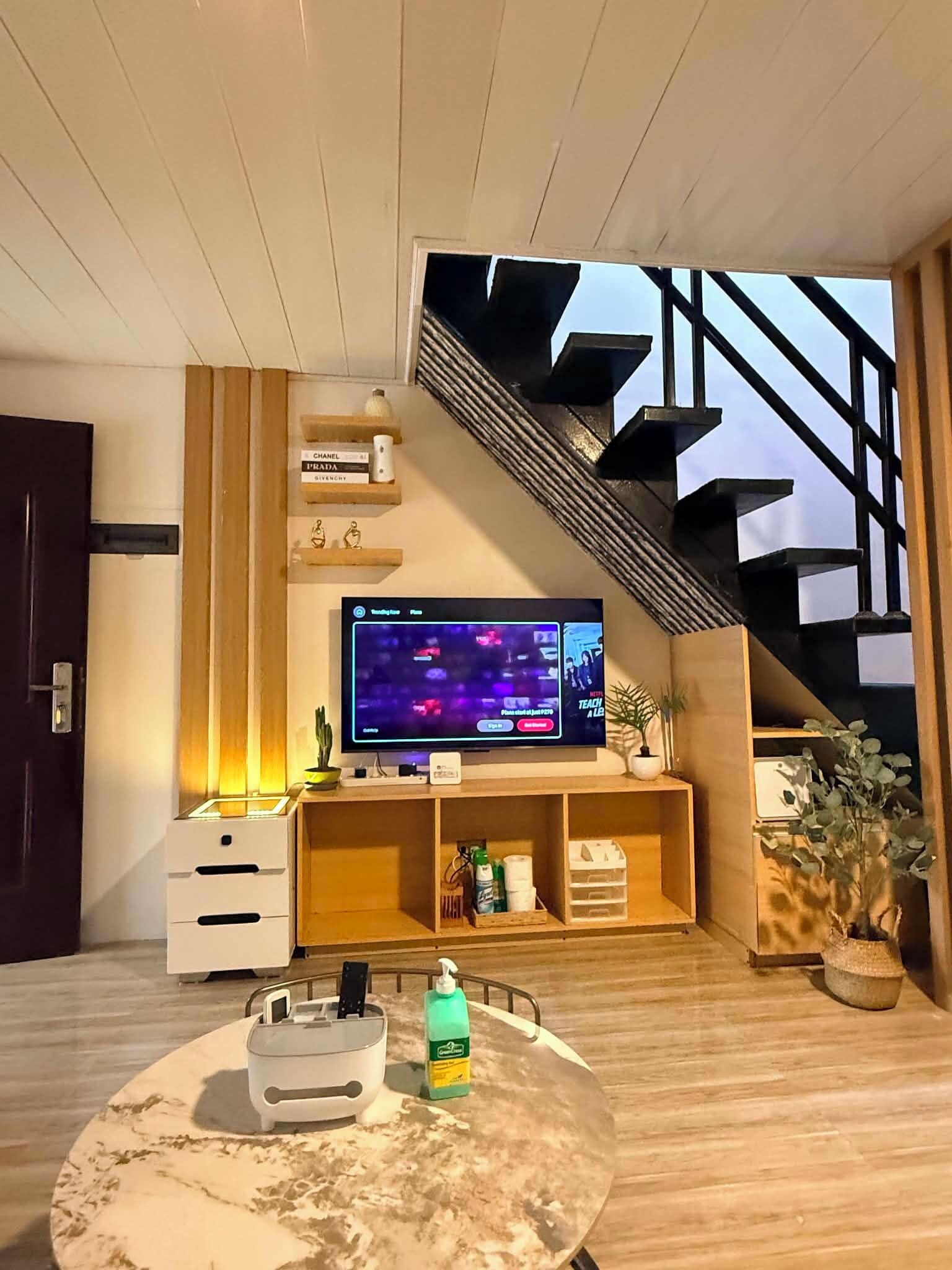 A modern living room with a flat-screen TV, a marble coffee table, a staircase, and a potted plant.