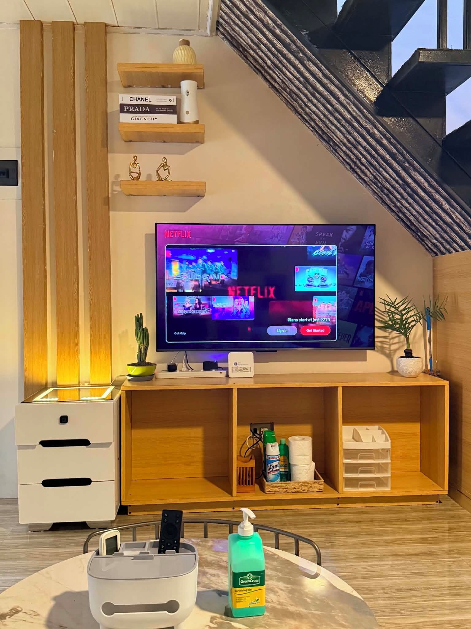 A modern living room with a flat-screen TV displaying a streaming service interface, wooden shelving with books, a small plant, and a remote control on a table.