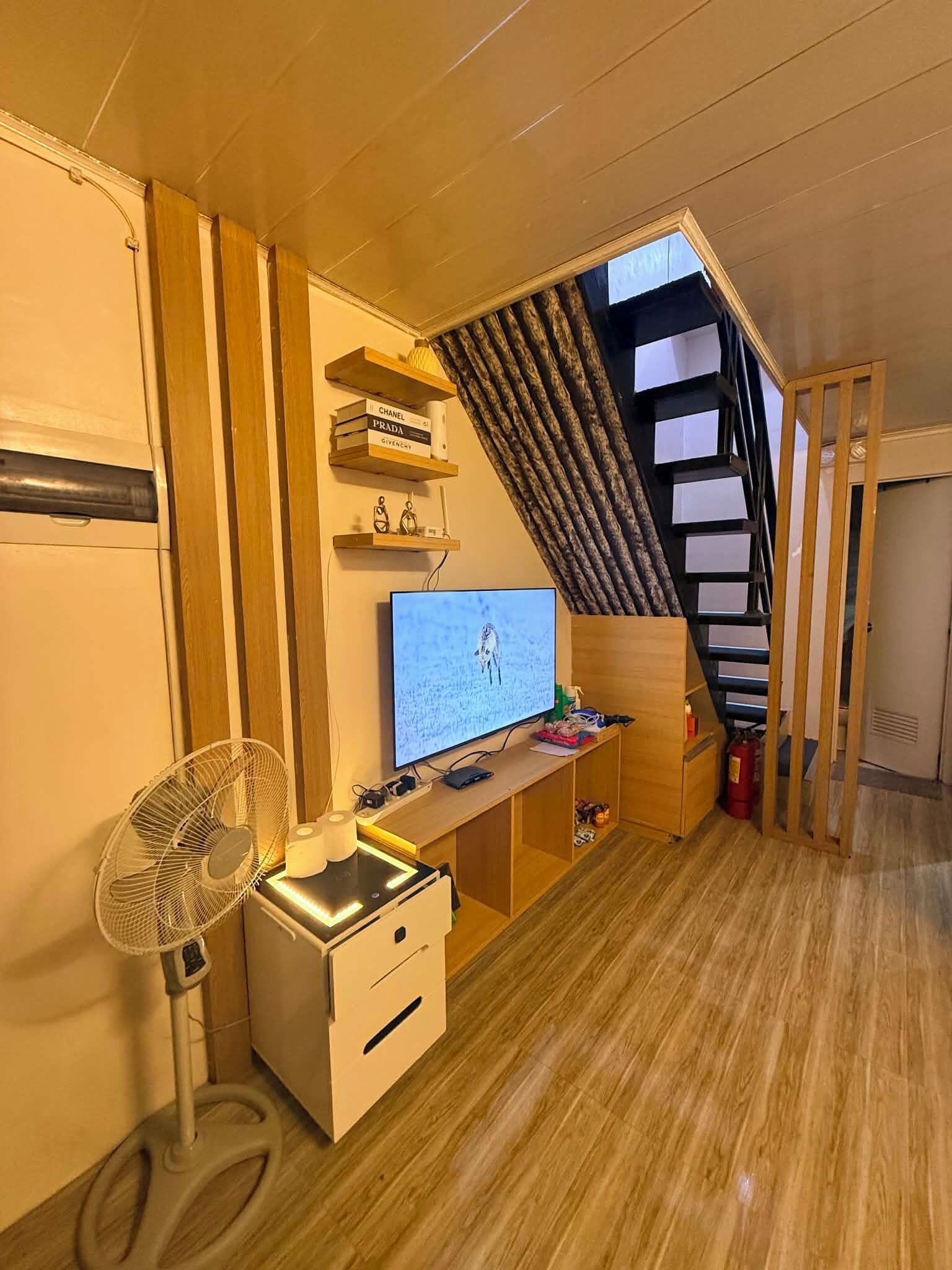 A compact room with a desk, a computer monitor, a fan, and a staircase.