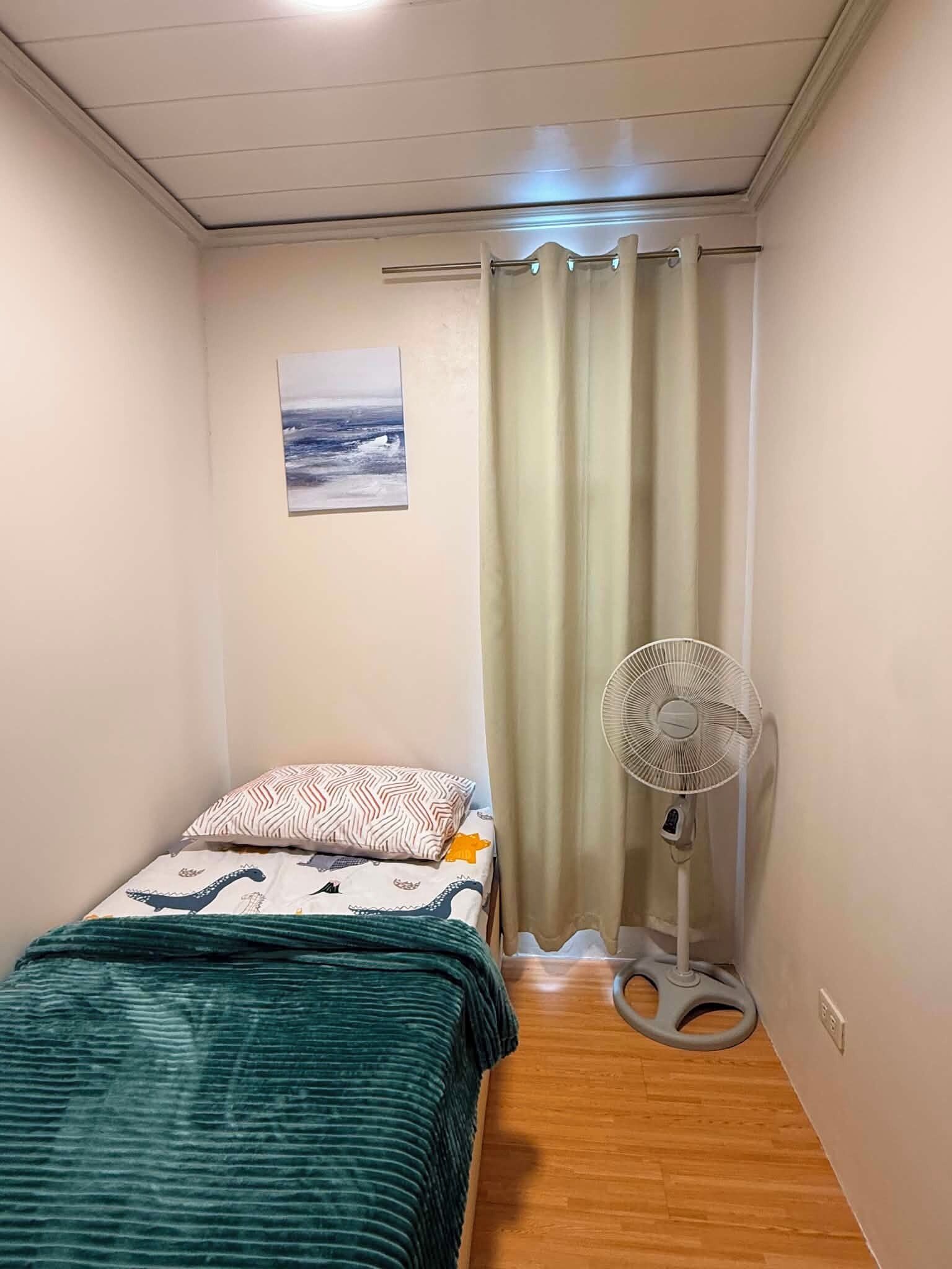 Une petite chambre avec un lit, un ventilateur sur pied, un rideau et un tableau accroché au mur.