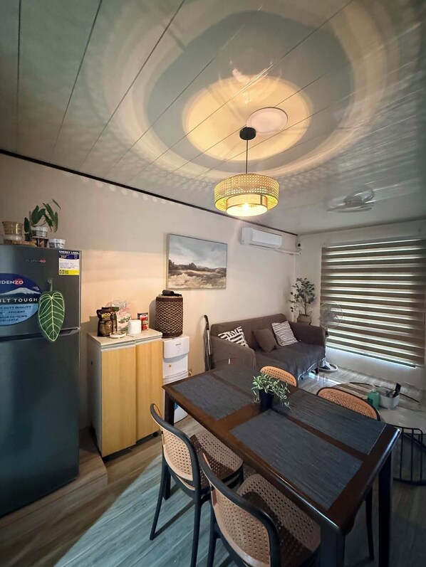 A modern living space with a dining table, chairs, a sofa, a refrigerator, and a ceiling light fixture.