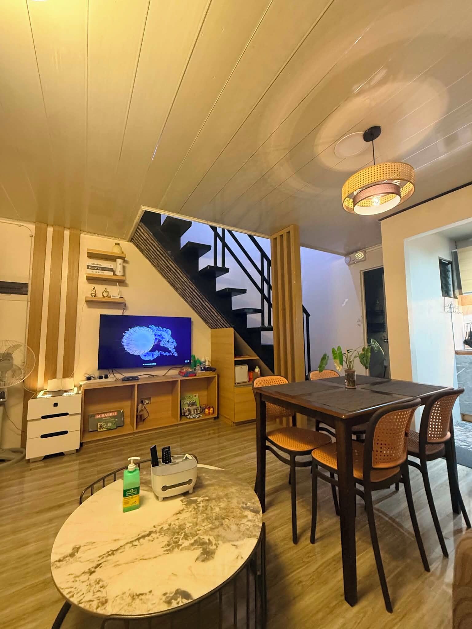 A modern living room with a wooden staircase, a dining table with chairs, a television, and a fan.