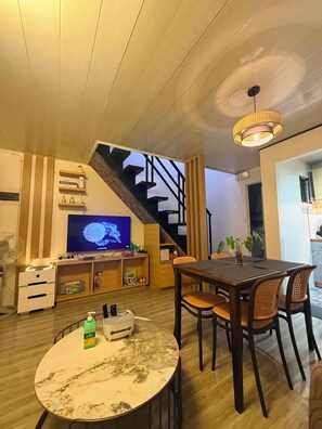 A modern living room with a wooden staircase, a dining table with chairs, a television, and a fan. - Balay Navona Matina l 3BR 2 Story Townhouse • Near SM Davao City (Davao City)