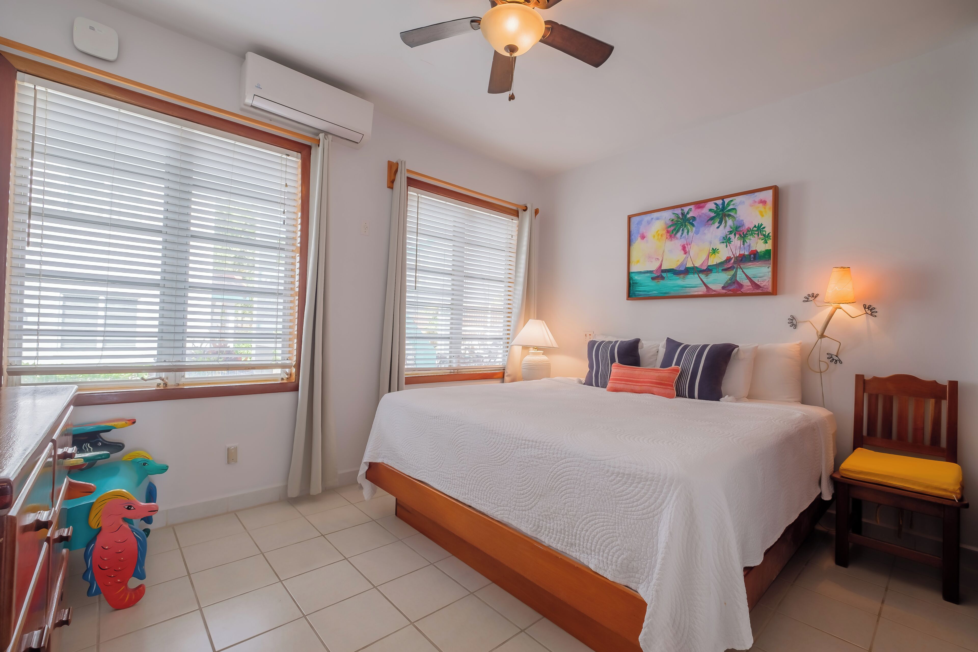 Quarto com cama, duas janelas com persianas, ventilador de teto, quadro, abajur e uma cadeira.