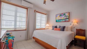 A bedroom with a bed, two windows with blinds, a ceiling fan, a painting, a lamp, and a chair.