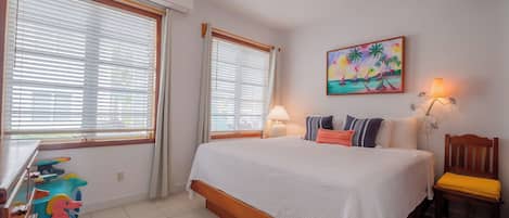 Un dormitorio con una cama, dos ventanas con persianas, un ventilador de techo, un cuadro, una lámpara y una silla.