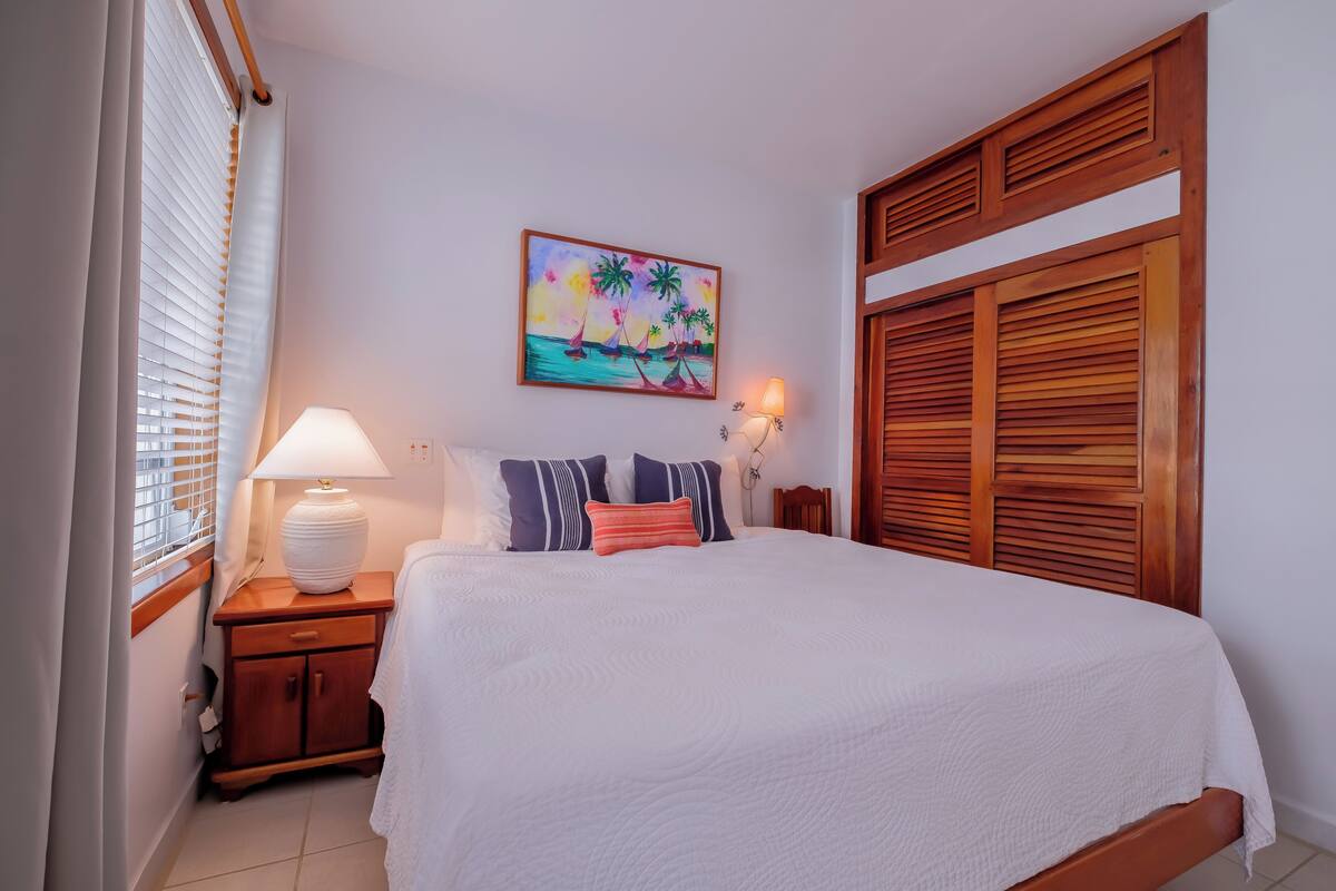 Una cama bien hecha con una colcha blanca, un cabecero de madera y un cuadro con una escena tropical encima.