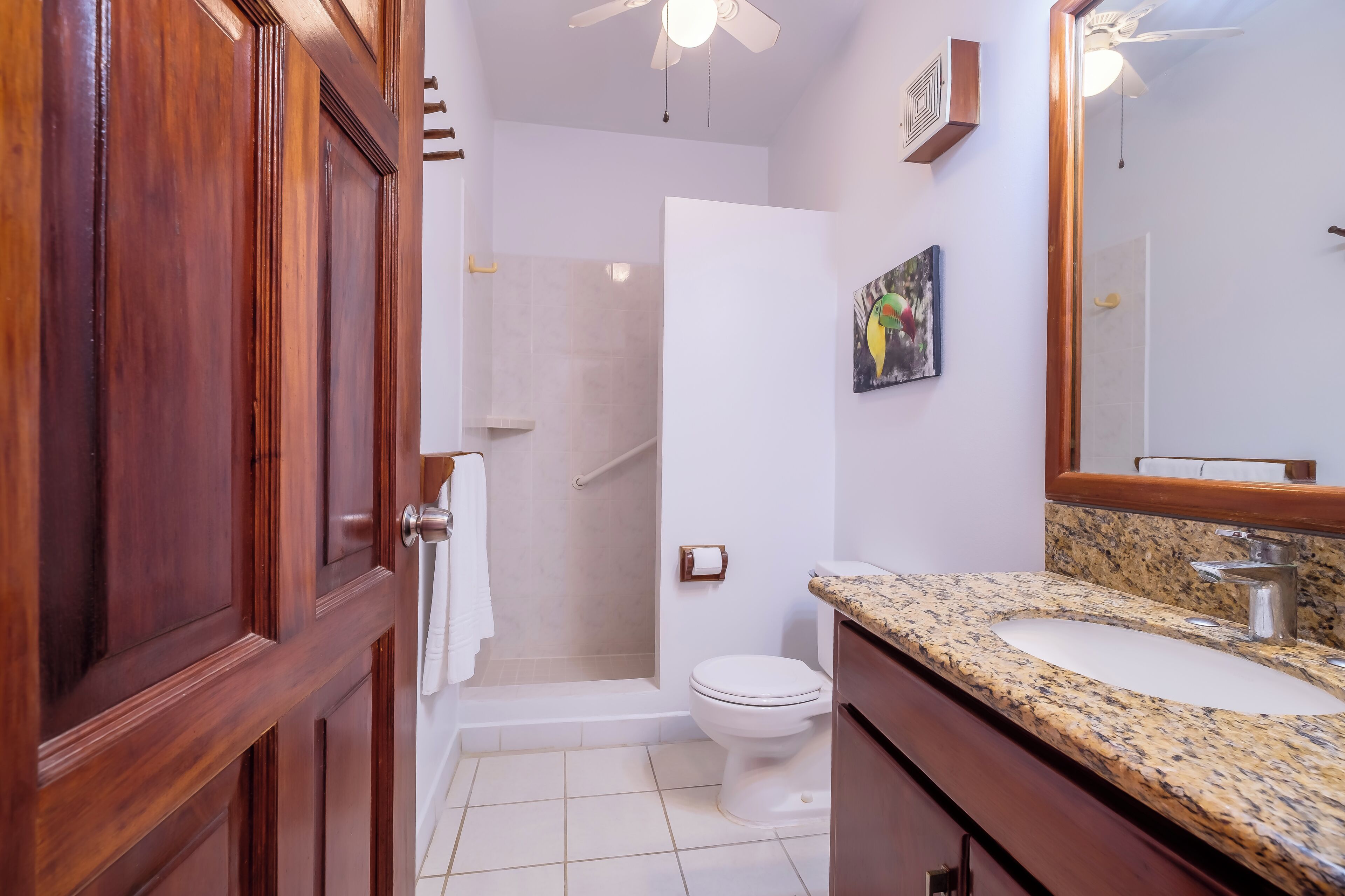 Un baño con puerta de madera, ducha, inodoro, lavamanos con encimera de granito y espejo.