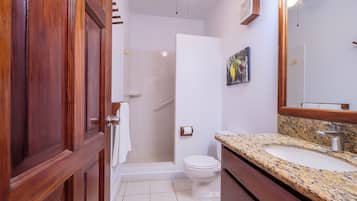 Un baño con puerta de madera, ducha, inodoro, lavamanos con encimera de granito y espejo.