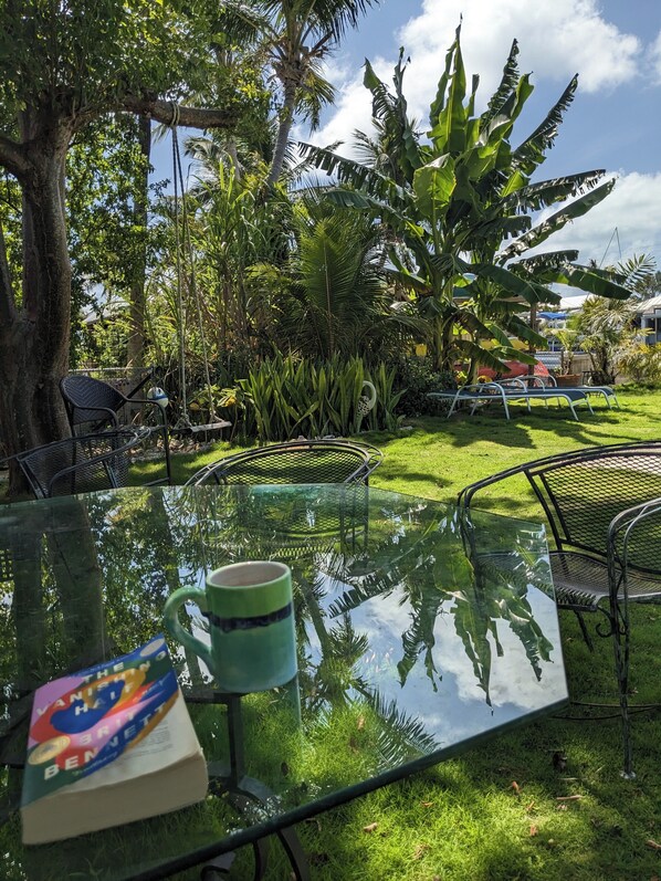 Une table en verre, sur laquelle reposent un livre et une tasse, entourée de chaises et d’une végétation luxuriante.