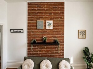 Ein Zimmer mit einer Ziegelwand, einem schwarzen Regal mit Topfpflanzen, einem grünen Sofa mit runden Kissen und gerahmten Kunstwerken.