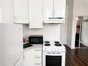 A kitchen with white cabinets, a black microwave, a stove with four burners, and a refrigerator. - Cozy 1 Bedroom in the heart of downtown Mtl (Montréal)