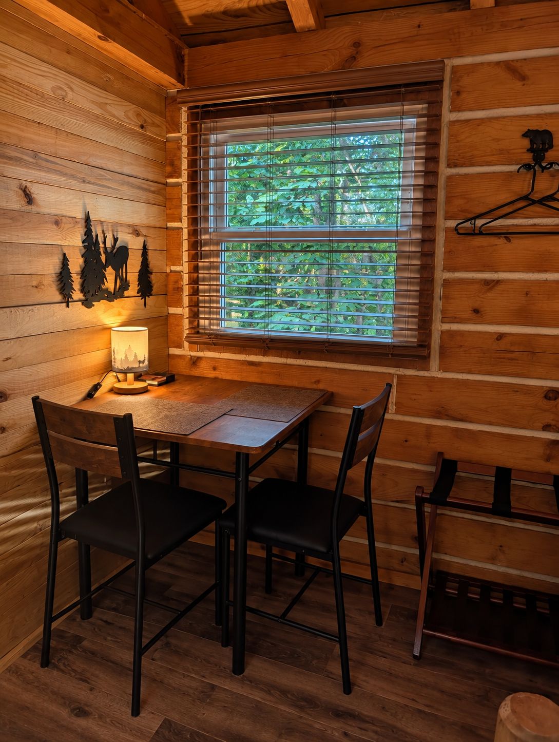 L'intérieur d'un chalet en bois comprend une table à manger, deux chaises, une fenêtre avec des stores et une lampe fixée au mur.