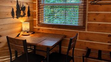L'intérieur d'un chalet en bois comprend une table à manger, deux chaises, une fenêtre avec des stores et une lampe fixée au mur.